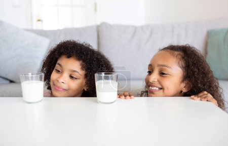 Photo for Lunch Drink. Happy Black Kid Girls Looking At Glasses With Milk On Table Posing At Home. Preteen Siblings Enjoying Healthy Beverages In Cozy Domestic Interior. Children Nutrition Concept - Royalty Free Image