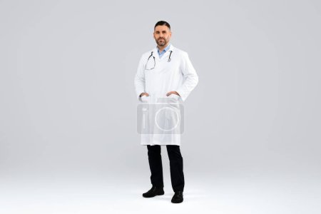 Photo for Confident middle aged doctor in white workwear looking at camera, posing with hands in pockets on light studio background, full length. Healthcare, medical, clinic concept - Royalty Free Image