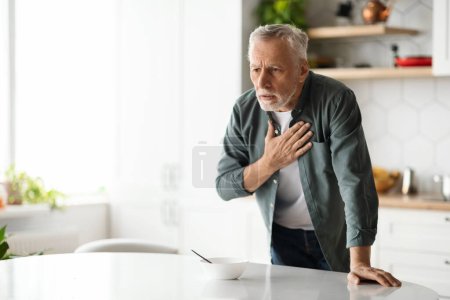 Heart Attack. Senior man suffering from chest pain at home, elderly gentleman having cardiac illness, feeling unwell, standing at table in kitchen interior and rubbing thorax area, copy space