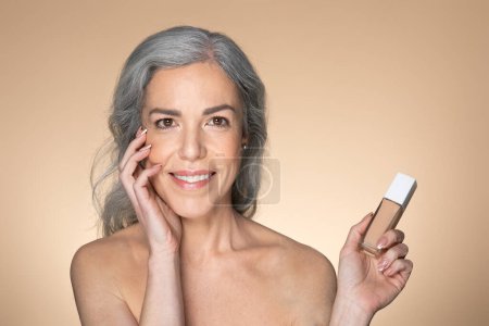 Téléchargez les photos : Portrait de belle femme européenne âgée à la peau claire et éclatante, tenant une bouteille de maquillage de fond de teint, posant sur un fond de studio beige. Recommandation de produit de maquillage - en image libre de droit