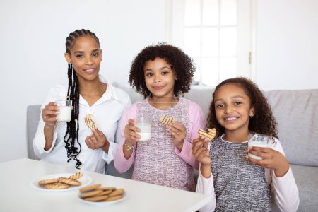 Photo for Snack Time. Positive Black Family Of Mother And Two Preteen Daughters Having Lunch Together, Eating Cookies And Drinking Milk At Home, Smiling To Camera. Mom And Children Posing Enjoying Food - Royalty Free Image