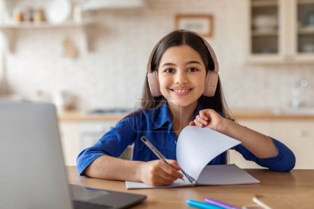 Foto de E-Learning fácil. Chica de la escuela feliz escribiendo en libro de texto estudiando en el ordenador portátil, posando en el escritorio en casa, sonriendo a la cámara. Niño usando auriculares y tomando notas aprendiendo en línea a través de la computadora - Imagen libre de derechos