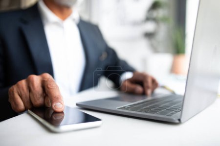 Photo for European senior businessman in suit sits at table with computer, typing on smartphone with blank screen in office interior. App for work, business remotely, blog, website and device - Royalty Free Image