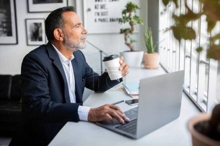 Téléchargez les photos : Heureux cher homme d'affaires européen senior en costume assis à la table à l'ordinateur, boire une tasse de café à emporter, pense, profiter de créer des idées et des solutions dans l'intérieur du bureau. Pause au travail, affaires - en image libre de droit