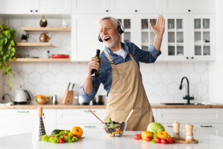 Téléchargez les photos : Homme âgé joyeux s'amusant tout en cuisinant dans la cuisine, en chantant et en dansant, Gentleman âgé heureux dans les écouteurs sans fil en utilisant la bouteille avec de l'huile d'olive comme microphone, préparer le déjeuner à la maison - en image libre de droit