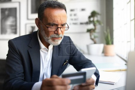 Photo for Concentrated european senior businessman in suit, glasses use smartphone, credit card at table in office interior. Problems at finance, business remotely, money banking app - Royalty Free Image