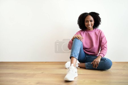 Téléchargez les photos : Joyeux millénaire afro-américain dame bouclée dans casual assis sur le sol dans le salon intérieur, sur le mur blanc. Reposez-vous, détendez-vous, pause et mode de vie à la maison seul, annonce et offre - en image libre de droit