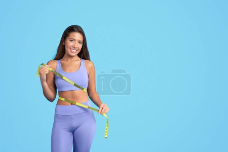 Photo for Smiling millennial caucasian slim woman in sportswear checking waist with measuring tape, isolated on blue studio background. Sports, weight loss result and body care, diet, ad and offer - Royalty Free Image