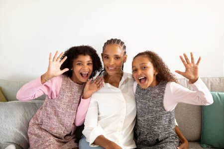 Foto de ¿Hola? Divertidas y juguetonas hijas afroamericanas y su madre saludando con las manos, posando y divirtiéndose sentadas en el sofá en casa. Familia de tres sonriendo a cámara gestos hola juntos - Imagen libre de derechos