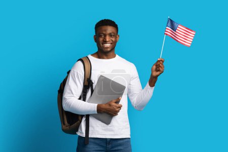 Téléchargez les photos : Joyeux étudiant millénaire afro-américain beau gars avec sac à dos tenant ordinateur portable et drapeau américain sur fond de studio bleu. Éducation à l'étranger, visa étudiant pour les États-Unis concept - en image libre de droit