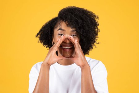 Photo for Cheerful millennial african american curly woman in white t-shirt presses hands to mouth, talk, shouts, isolated on yellow studio background, close up. Lifestyle, secret, great news, ad and offer - Royalty Free Image