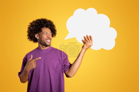 Téléchargez les photos : Homme bouclé adulte noir joyeux en t-shirt violet pointe du doigt un nuage abstrait avec un espace vide, isolé sur fond orange, studio. Recommandation, conseils, vente et pensées, annonce et offre - en image libre de droit