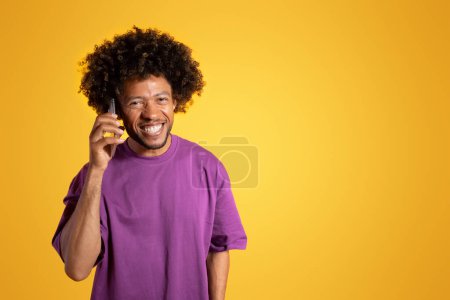Photo for Positive black adult curly man in purple t-shirt calls by smartphone, talks isolated on orange background, studio. Great news, gossip, communication remotely, ad and offer - Royalty Free Image