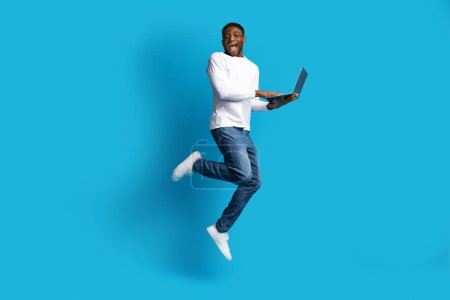 Photo for Amazing online offer. Happy excited young black guy in casual outfit jumping in the air with modern pc laptop in his hands over blue studio background, full length, copy space - Royalty Free Image