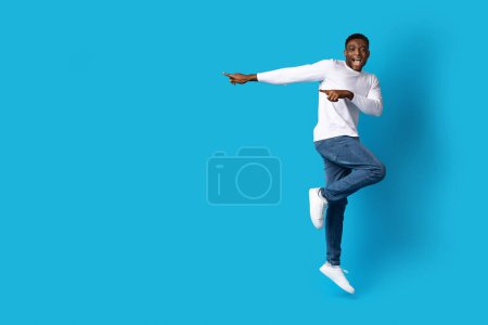 Photo for Nice offer. Amazed happy handsome young black guy in casual hipster jumping in the air over blue studio background, pointing at copy space for text or advertisement, full length - Royalty Free Image