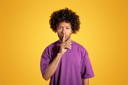 Photo for Serious black adult curly man in purple t-shirt presses finger to lips, making shh sign, isolated on orange background, studio. Secret news, surprise gesture, ad and offer - Royalty Free Image