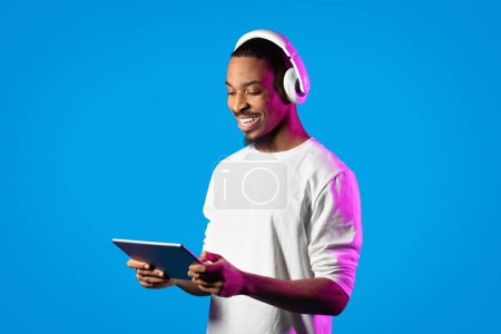 Photo for Entertainment and modern technologies. Happy attractive young african american guy in white shirt using modern digital tablet and wireless headphones in neon light over blue background, copy space - Royalty Free Image