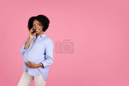 Foto de Feliz joven embarazada afroamericana rizada dama en casual con gran vientre llamando por teléfono inteligente, aislado en fondo de estudio rosa. Espere bebé, comunicación, buenas noticias, chismes, anuncios y ofertas - Imagen libre de derechos