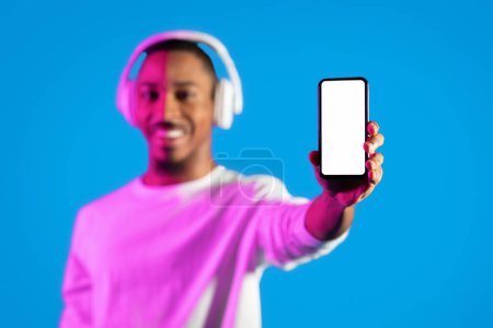 Téléchargez les photos : Smartphone avec écran blanc vide dans la main du mec afro-américain, joyeux jeune homme noir en utilisant des écouteurs sans fil montrant le téléphone, recommandant une belle application musicale mobile, fond bleu, maquette - en image libre de droit