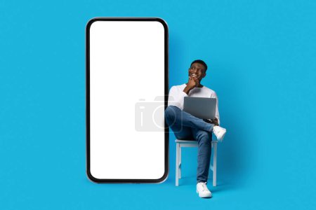 Photo for Excited curious young black man in casual outfit independent contractor sitting on chair with computer on his lap, looking at smartphone with white blank screen, mockup for booking mobile app - Royalty Free Image