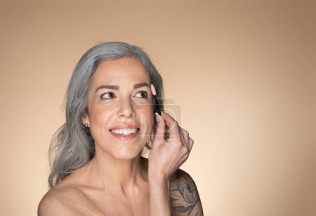 Glückliche grauhaarige Seniorin, die sich die Augenbrauen putzt, Pinsel hält und lächelt, Schönheitsroutine und Kosmetikprodukt genießt, auf beigem Hintergrund posiert, freier Raum