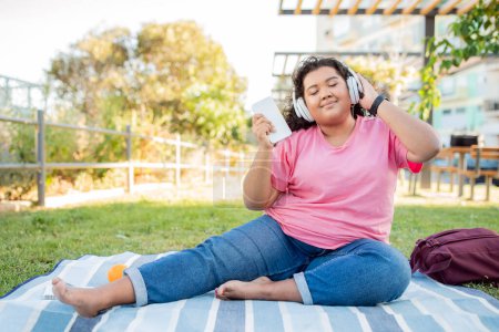 Téléchargez les photos : Happy Overweight Latin Woman Écouter de la musique en ligne avec des écouteurs et un smartphone, assis sur une couverture profiter du pique-nique dans le parc à l'extérieur. Lady Closing Eyes, Écoute ma playlist préférée le jour de l'été - en image libre de droit