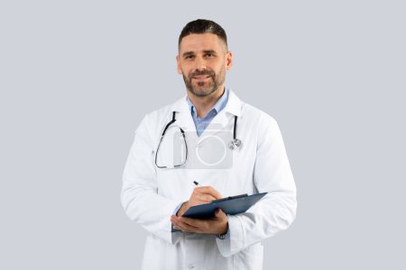 Photo for Professional doctor man in medical coat with clipboard in hands posing over light grey wall background, middle aged physician with stethoscope looking at camera - Royalty Free Image