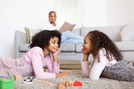 Photo for Siblings Pastime. Cheerful Black Preteen Sisters Girls Having Fun Playing With Toys Together While Mom Reading A Book In Cozy Living Room At Home. Selective Focus On Children - Royalty Free Image