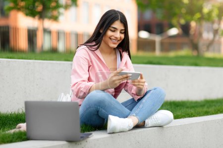Téléchargez les photos : Heureuse dame hispanique en utilisant un téléphone portable jouer à des jeux et apprendre en ligne, assis sur le campus ou le parc de l'université. Gadgets concept de loisirs et d'enseignement à distance - en image libre de droit