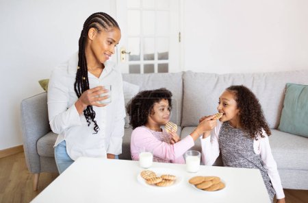 Photo for Family Lunch. Happy Black Mommy Looking At Her Kids Daughters While They Having Fun Eating Cookies And Drinking Milk, Feeding Each Other At Table At Home. Healthy Nutrition For Children Concept - Royalty Free Image