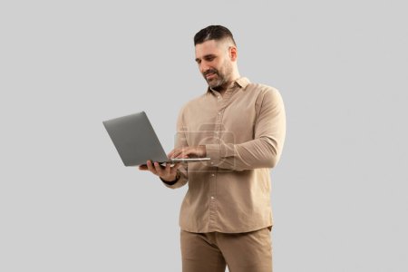 Photo for Positive middle aged caucasian man typing on laptop computer, surfing in internet, working online, standing on light studio background. Device for business, work remotely - Royalty Free Image