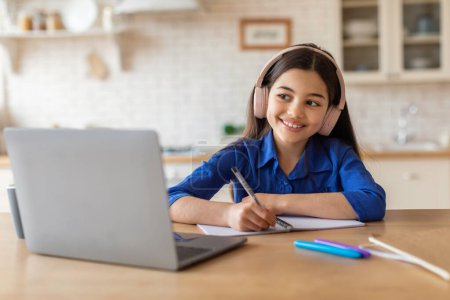 Foto de Oferta de E-Learning. Chica alegre del niño de la escuela que hace la tarea en el ordenador portátil, usando auriculares inalámbricos y tomando notas en casa. Colegiala aprendiendo a ver lecciones virtuales en la computadora. Estudio y Gadgets - Imagen libre de derechos