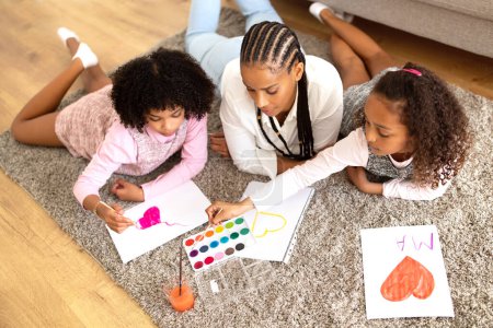 Photo for African American Mom And Two Daughters Drawing Together In Modern Living Room, High Angle View. Family Of Three Engaging in Painting Fun Making Greeting Postcard Together On Weekend - Royalty Free Image