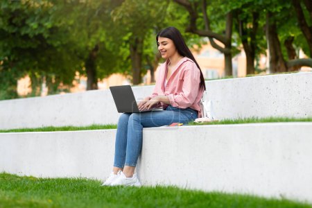Foto de Feliz adolescente hispana dama con portátil sentado en el parque, estudiante niña estudiando remotamente con la computadora al aire libre, escribiendo en el teclado y sonriendo, espacio de copia. Concepto de aprendizaje online - Imagen libre de derechos