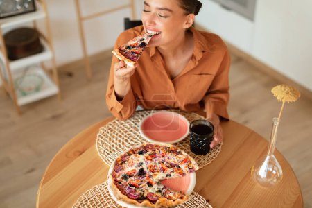 Photo for Happy caucasian woman biting a slice of pizza, enjoying tasty lunch at home, sitting at table in kitchen interior, above view. Cheerful lady having her meal alone - Royalty Free Image
