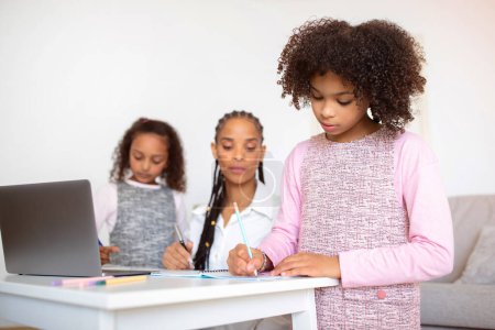 Photo for Domestic E-Learning. Black Preteen Girl Learning At Laptop And Taking Notes, While Mom And Little Sister Helping Her With Online Homework At Home. Modern Distance Education Concept - Royalty Free Image