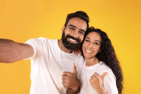 Téléchargez les photos : Nous aimons cette offre. Joyeux couple moyen-oriental faisant Selfie Gesturing pouces en haut dans la pose d'approbation sur fond jaune studio, souriant à la caméra. Conjoints approuvant quelque chose - en image libre de droit