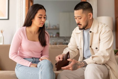 Finance Hardship And Povertry. Unhappy Young Asian Couple Looking At Empty Wallet With No Money, Struggling Financial Issues Sitting On Sofa At Home. Family Suffering From Economic Crisis