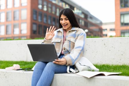 Aufgeregte Studentin winkt mit der Hand zum Laptop, während sie im Park der modernen Universität Online-Unterricht hat, Videotelefonie mit dem Lehrer. Fernstudium und E-Learning-Konzept