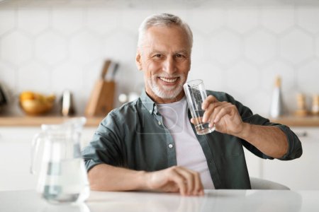 Foto de Caballero mayor feliz sosteniendo el vaso con agua mientras está sentado en la mesa en la cocina, sonriendo anciano disfrutando refrescante bebida y mirando a la cámara, hombre sediento tomando líquido saludable - Imagen libre de derechos