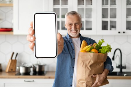 Photo for Grocery Delivery App. Smiling Senior Man Showing Blank Smartphone And Holding Paper Bag With Groceries, Happy Elderly Male Demonstrating Empty Cellphone, Advertising Application Or Website, Mockup - Royalty Free Image
