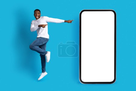 Photo for Positive happy excited young black guy wearing casual outfit jumping next to big phone over blue background, pointing at white blank screen, mockup for online offer or mobile app - Royalty Free Image