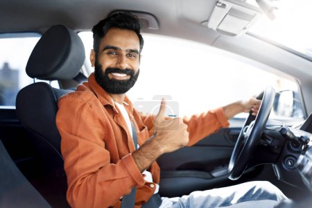 Foto de Auto nuevo. Happy Indian Driver Man Gesturing Thumbs Up Driving Car y sonriendo a la cámara sentado en el asiento sosteniendo las manos en el volante en el automóvil en interiores. Propiedad y transporte de vehículos - Imagen libre de derechos