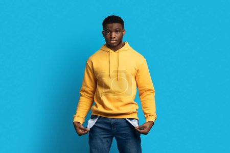 Photo for Poverty and absence of money during crisis. Unemployed jobless unhappy young african american man showing empty jeans pockets, blue studio background, copy space - Royalty Free Image