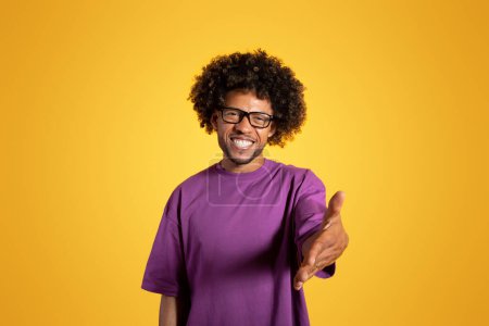 Photo for Happy black adult curly guy in violet t-shirt and glasses makes greeting gesture with hand, isolated on orange background, studio. Man gives hand to camera, say hello, ad and offer - Royalty Free Image