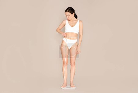 Photo for Slim slender beautiful caucasian young woman wearing white comfy underwear checking weight on floor scales and measuring her waist with tape, beige background, copy space, full length - Royalty Free Image