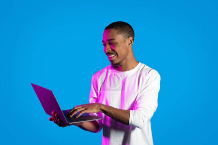 Téléchargez les photos : Freelance, concept de télétravail. Positif joyeux jeune homme afro-américain en blanc tapant sur le clavier de l'ordinateur portable pc et souriant, travaillant en ligne, isolé sur fond bleu dans la lumière au néon, espace de copie - en image libre de droit