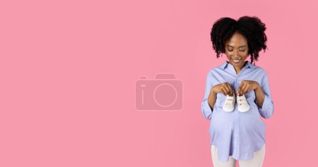 Photo for Happy young pregnant african american curly woman in casual puts little shoes on big belly, isolated on pink studio background, panorama. Expect baby, love, health care, advertising and offer - Royalty Free Image