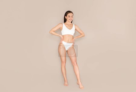 Photo for Cheerful pretty slender millennial woman wearing white comfortable underwear posing on beige studio background, demonstrating perfect body, beautiful curves, long fit legs, copy space - Royalty Free Image
