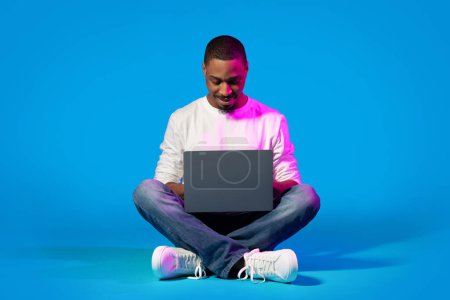 Téléchargez les photos : Joyeux sourire jeune homme élégant afro-américain beau assis sur le sol, en utilisant ordinateur portable sur fond bleu, websurfing, shopping en ligne, bavarder avec des dames en ligne, espace de copie - en image libre de droit
